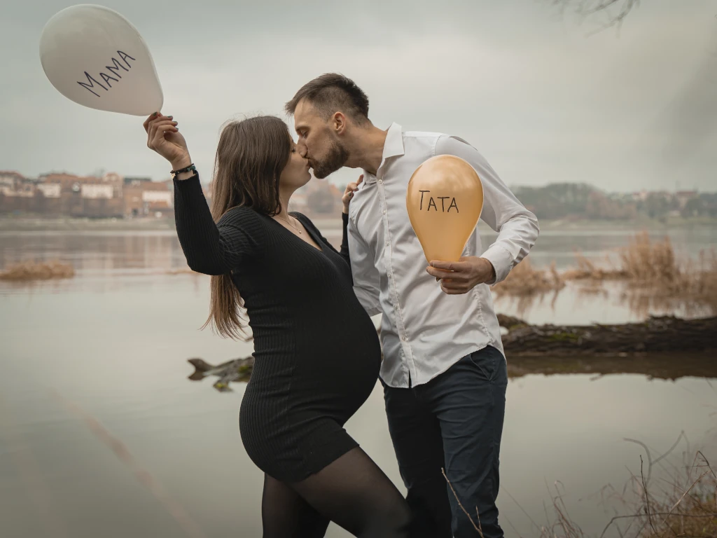 Plenerowa sesja ciążowa w Toruniu – para przyszłych rodziców całuje się nad Wisłą, trzymając balony z napisami Mama i Tata.