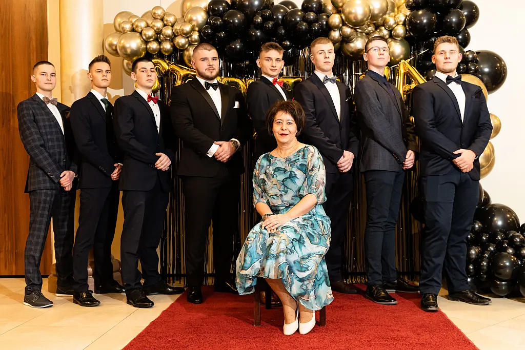 Studniówka 2023 Toruń – uczniowie w eleganckich garniturach z wychowawczynią na tle dekoracji balowej